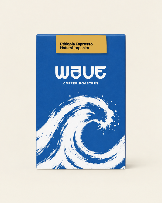 A rectangular coffee roaster box standing upright on a light beige background. The box has a blue or a dark blue design with a large white wave illustration and the visible brand name 'wave coffee roasters' with specialty coffee label: 'Ethiopia Espresso Natural (organic)'