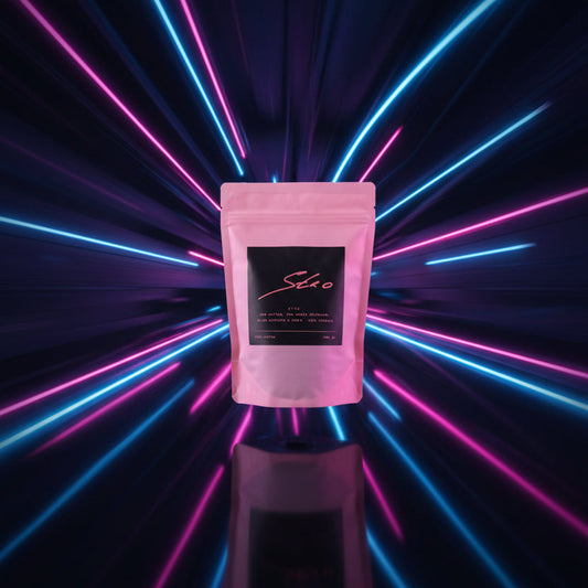 A pink stand-up pouch of stro coffee with a black label, standing on a white table surrounded by matching branded cards. The background shows a blurred retail or office interior with shelves and glass doors.