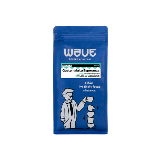 A blue stand-up pouch of Wave Coffee Roasters Guatemala La Esperanza medium roast coffee, featuring white text and a cartoon illustration of a man balancing coffee cups on a white background.