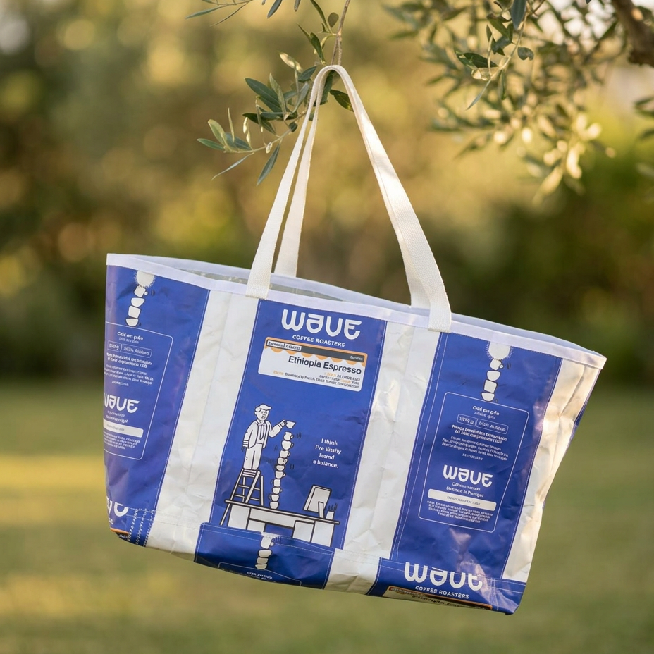 A reusable shopping tote bag made from blue and white plastic coffee packs, hanging from olive tree branches outdoors with a blurred green garden background.