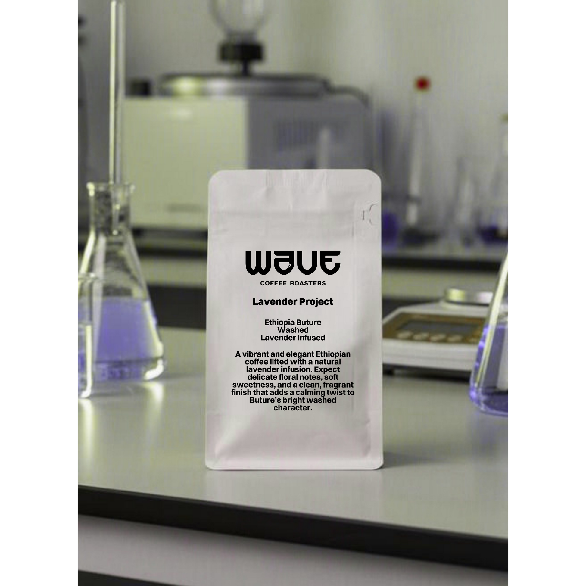 A white stand-up pouch of Wave Coffee Roasters 'Lavender Project' stands on a reflective countertop in a laboratory setting with beakers and glassware blurred in the background.